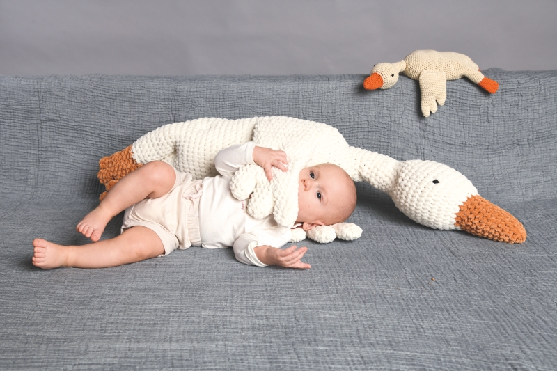 Baby in weißem Strampler mit handgemachtem Strickgans-Spielzeug. Modische, dezente Farbgestaltung.