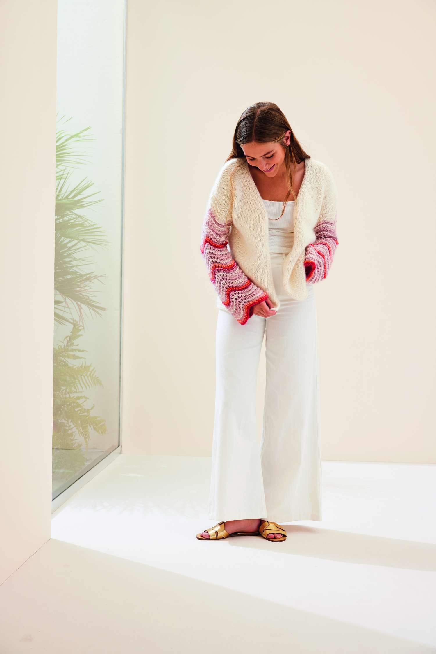 Weißer Cardigan mit rosa-rot gemusterten Ärmeln, kombiniert mit modischer weißer Hose, Goldsandalen.