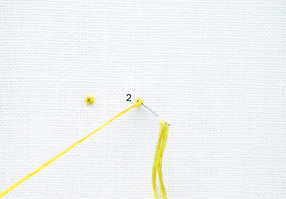 Gelber Faden bildet einen französischen Knoten auf weißem Stoff. Handarbeit, präzise Detailarbeit.