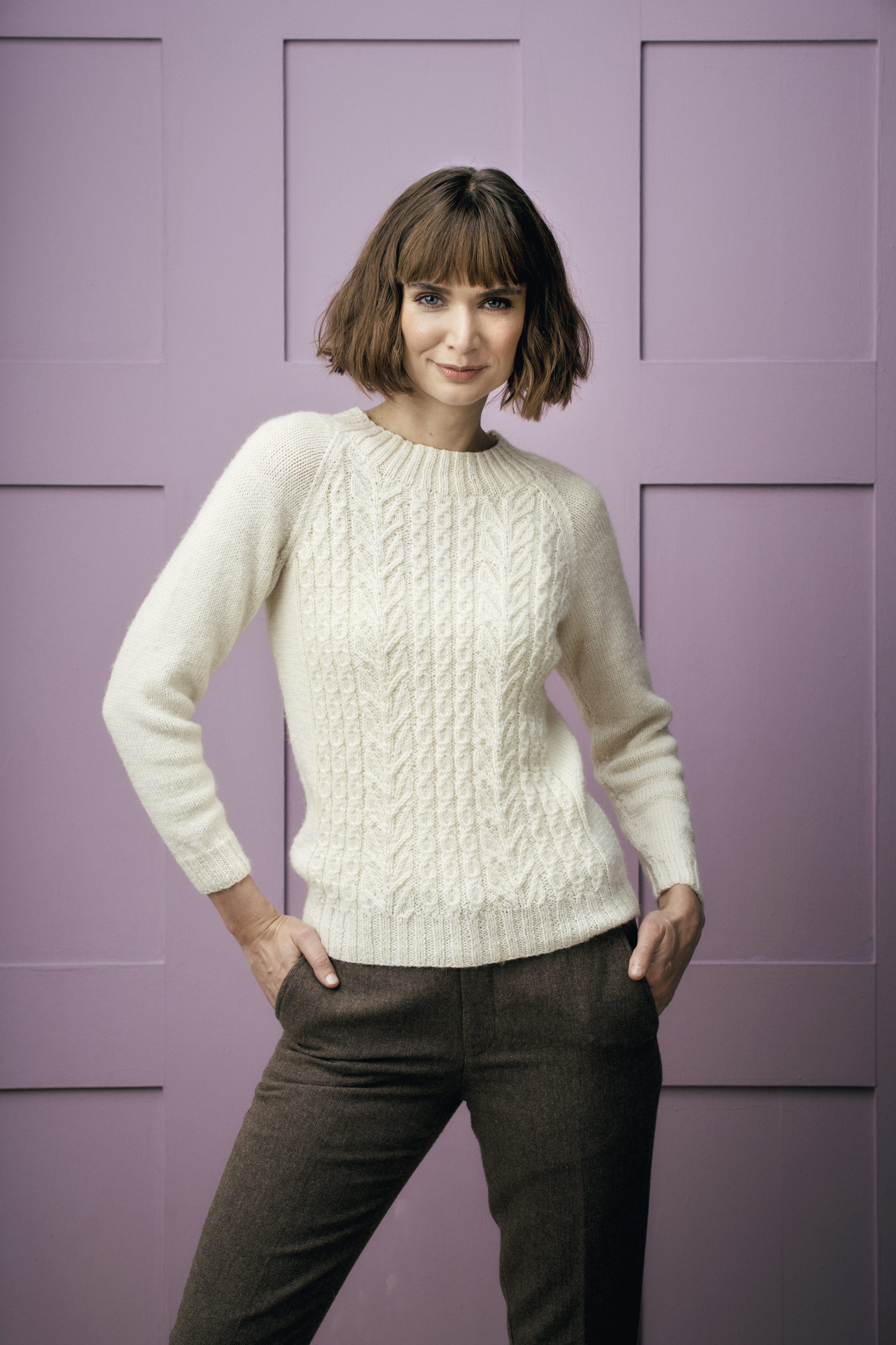 Weißer, modischer Strickpullover mit Zopfmuster, braune Hose, schlichter Schnitt. Handarbeit, elegant.