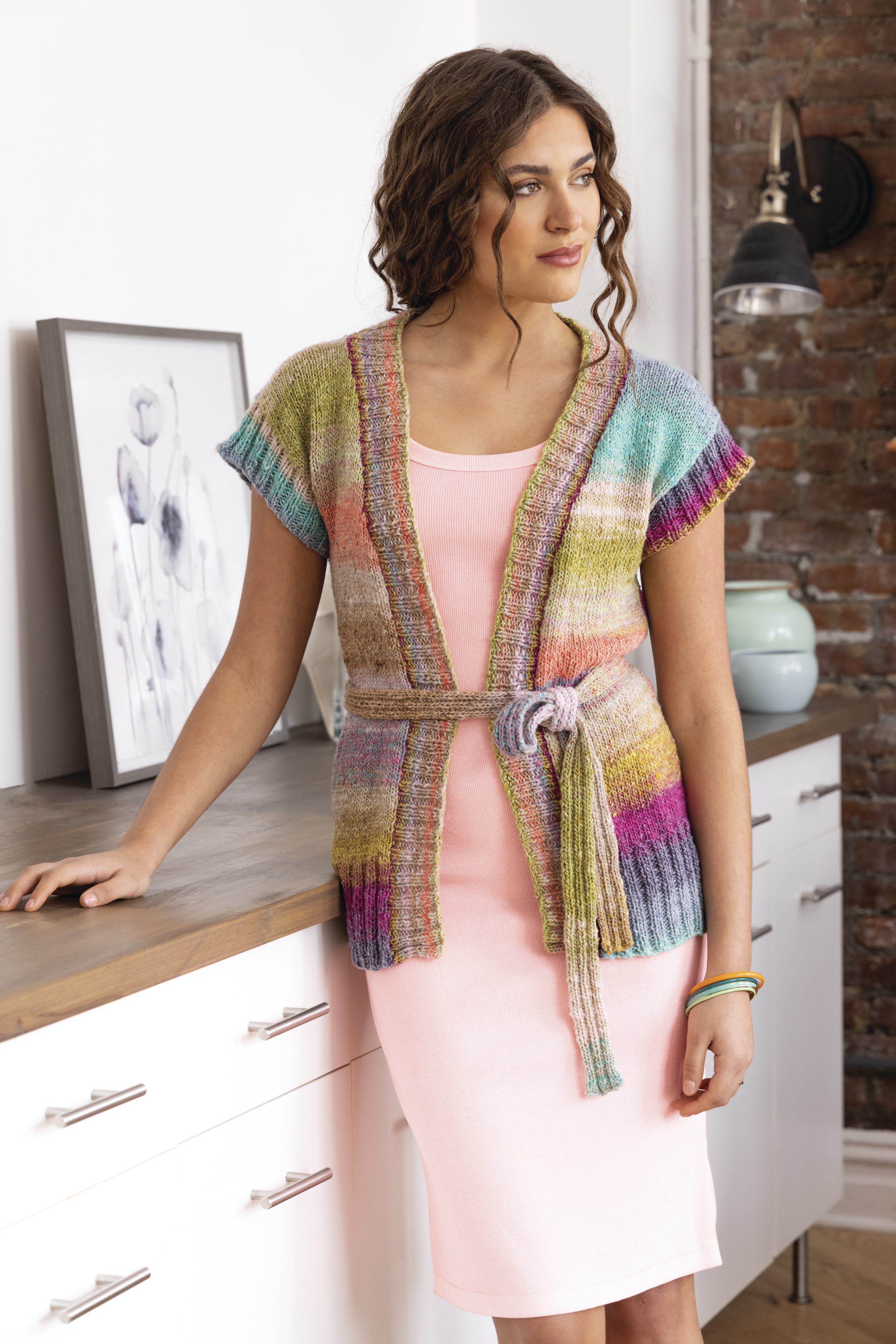 Bunter, handgemachter Cardigan mit Regenbogenstreifen, gebunden über pastellfarbenes Kleid. Modisch und einzigartig.
