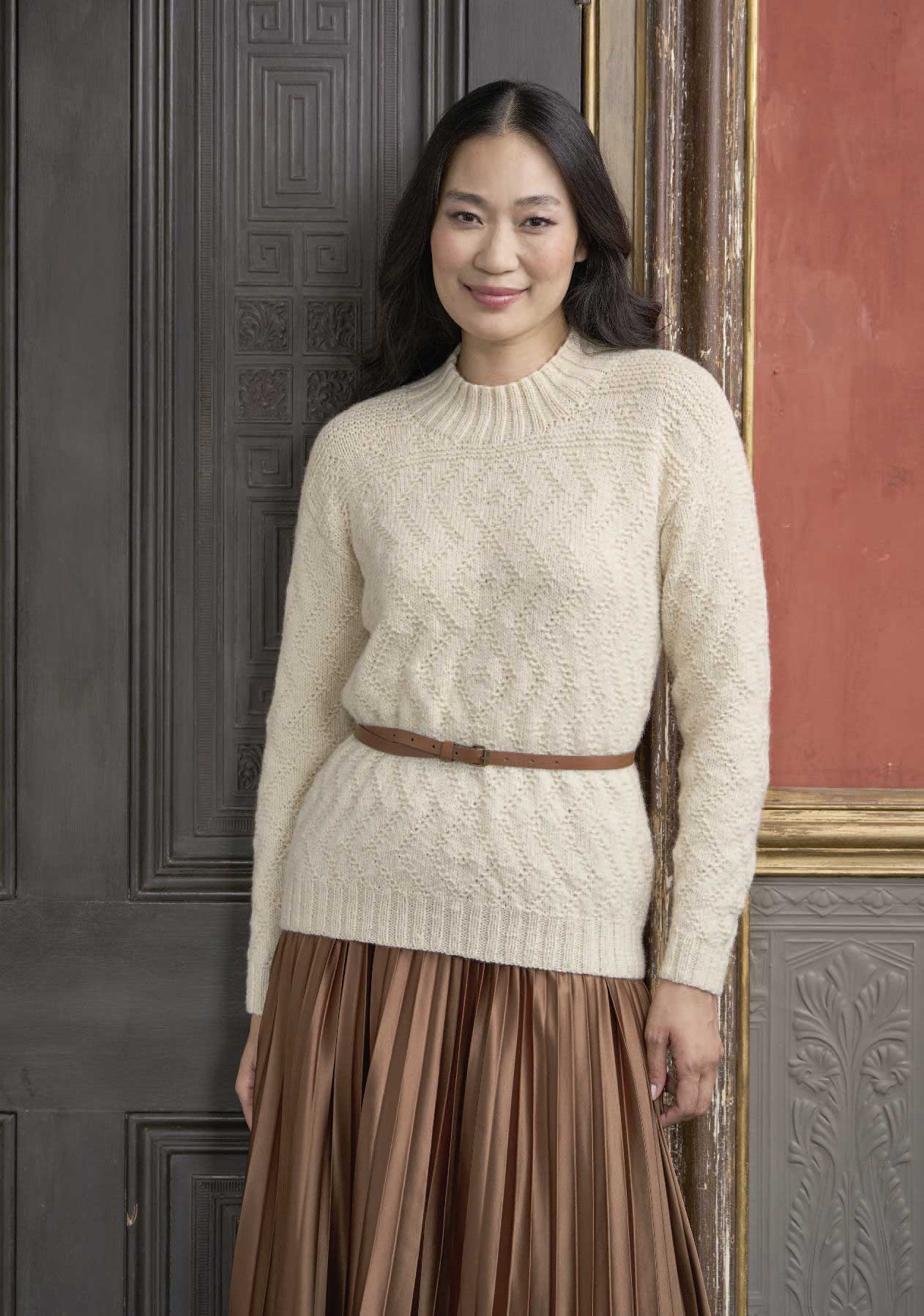 Cremefarbener Strickpullover mit Zopfmuster, brauner Ledergürtel, bronzefarbener Faltenrock. Modisch, handgemacht.