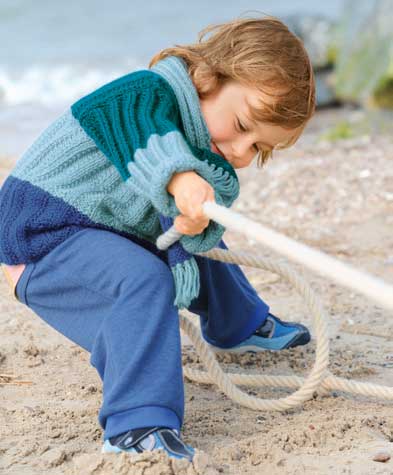 Kind trägt modischen, handgestrickten Pullover in Blaugrün mit Zopfstrickmuster; bequeme, blaue Hose. DIY-Chic.