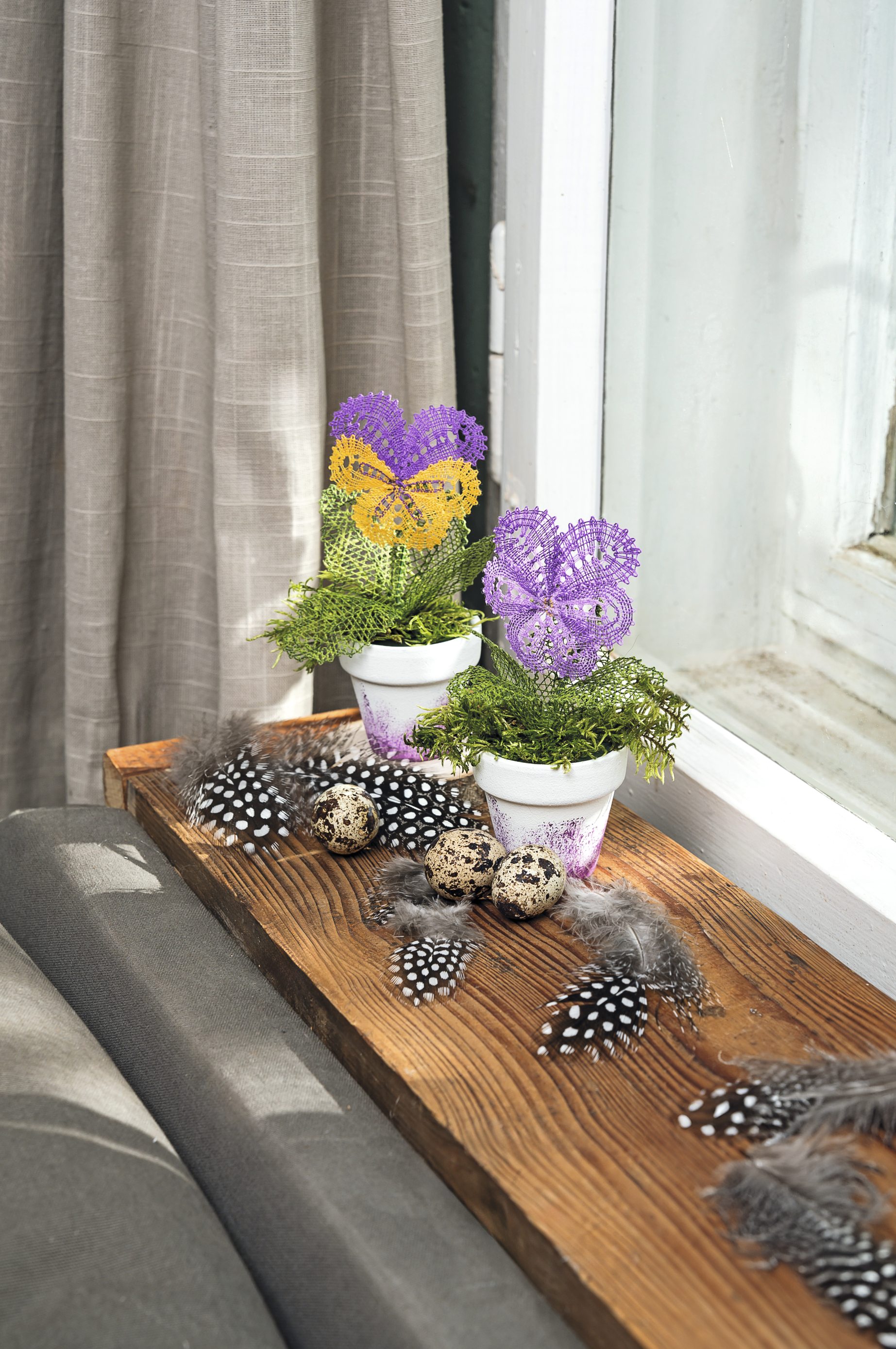 Bunte, selbstgemachte Häkelblüten in Töpfen, begleitet von Federn und Eiern für einen kreativen und natürlichen Look.