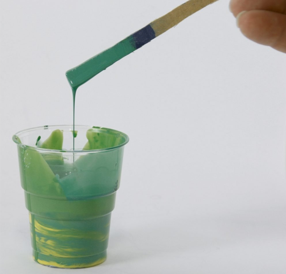Grüner Farbton tropft von einem Holzstab in einen durchsichtigen Becher mit gelbem Muster. DIY, kreativ, handgemacht.