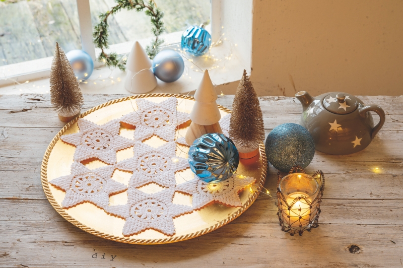 Handgemachte weiße Häkelsterne auf goldener Platte, umgeben von blauen Kugeln. DIY, festlich, elegant.