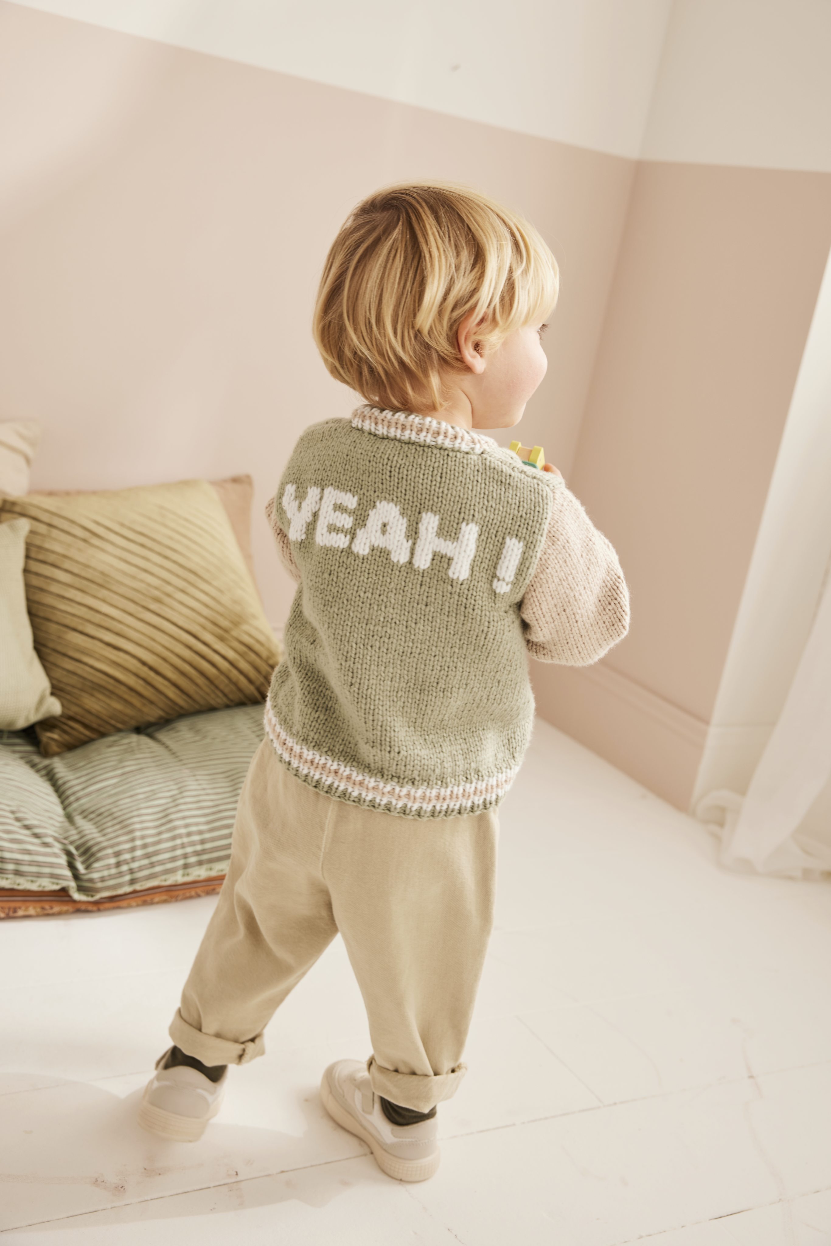 Selbstgemachter, pastellgrüner Strickpullover mit 'YEAH!' Muster und beigen Ärmel, kombiniert mit hellen Chinohosen.