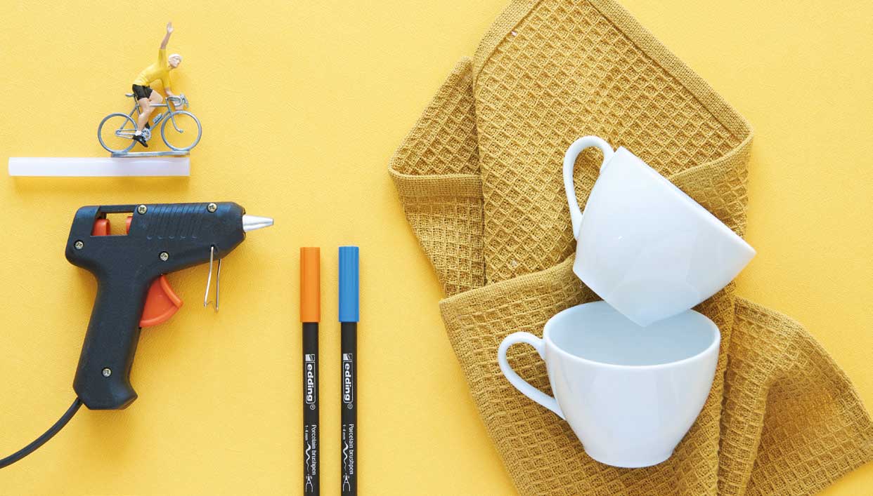 Farbenfrohes DIY-Arrangement: Heißklebepistole, Stifte, zwei weiße Tassen, strukturierter Stoff, Mini-Figur.
