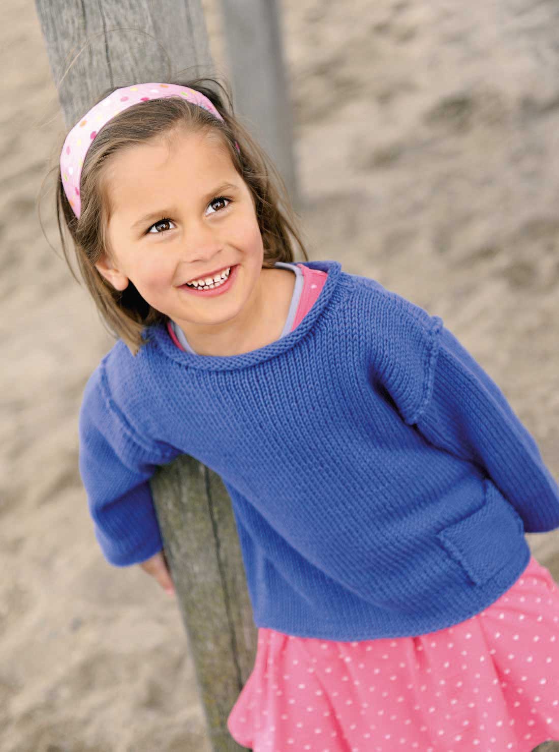 Kind trägt blauen Pullover, handgestrickt; pinkes Kleid mit weißen Punkten: verspielter, selbstgemachter Stil.