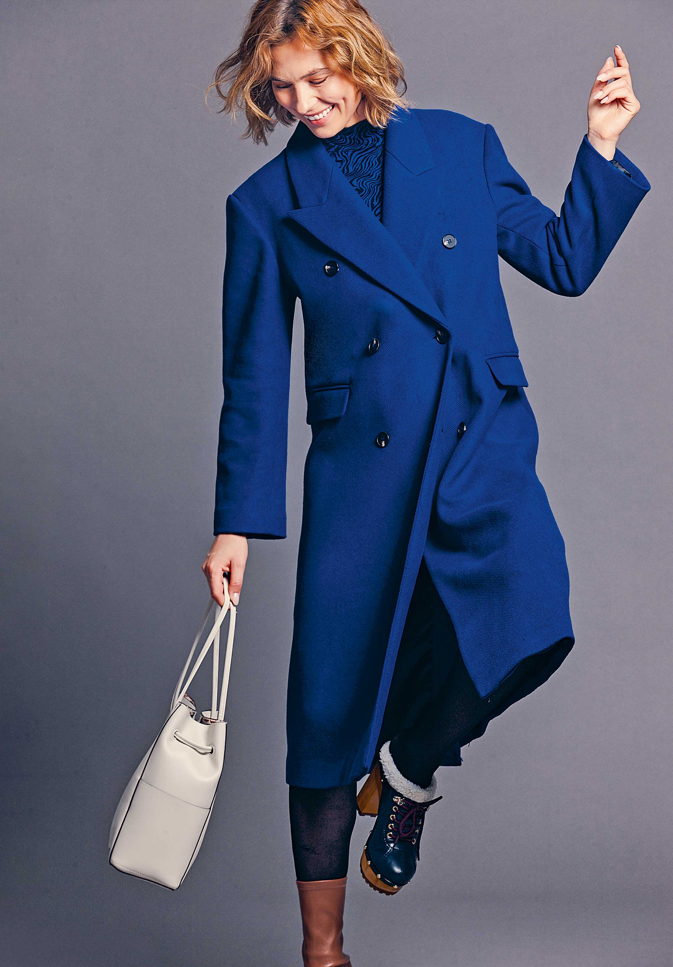 Modischer blauer Mantel, doppelreihig, elegant geschnitten. Dunkle Knöpfe, kombiniert mit cremefarbener Tasche.