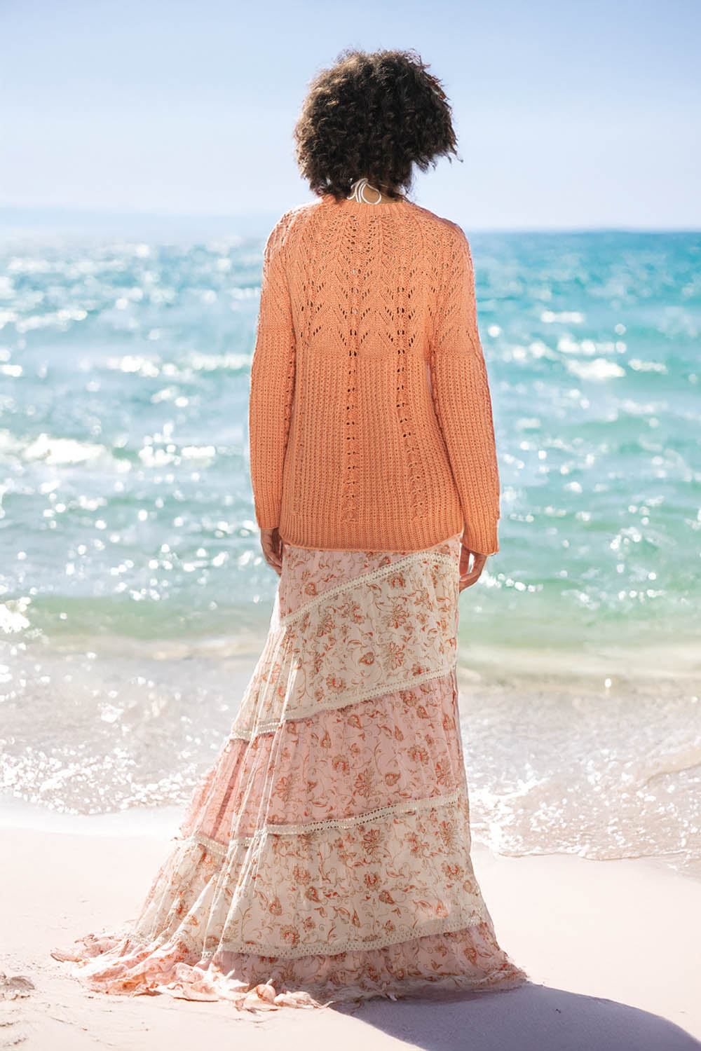 Orange Strickpulli, lang, kombiniert mit floralem Maxirock in Pastelltönen, modisch und leicht. Handarbeitsflair.