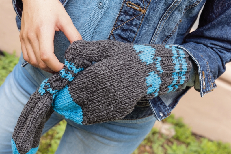 Grau-blaue strickte Fäustlinge, handgemacht, mit modischen Mustern. Kombiniert mit Jeansjacke und heller Jeans.