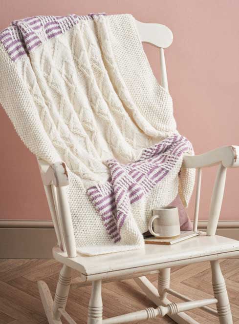 Weiße Strickdecke mit Zopfmuster und lila Karos, handgestrickt. Perfekt für gemütliche Abende.