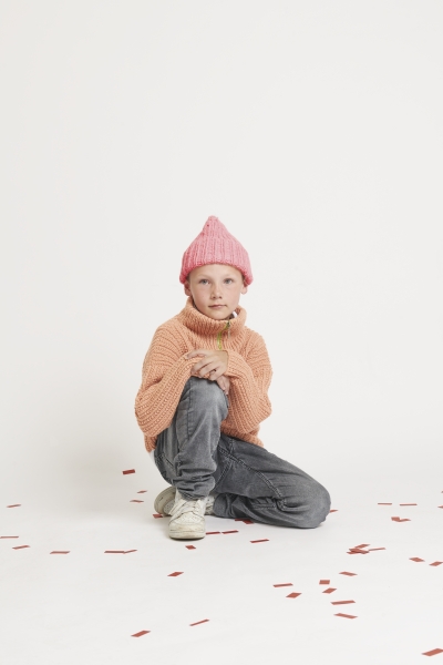 Kind in grobem, pfirsichfarbenem Strickpullover, rosa Mütze; modisch, handgemacht, mit lässigen Jeans.