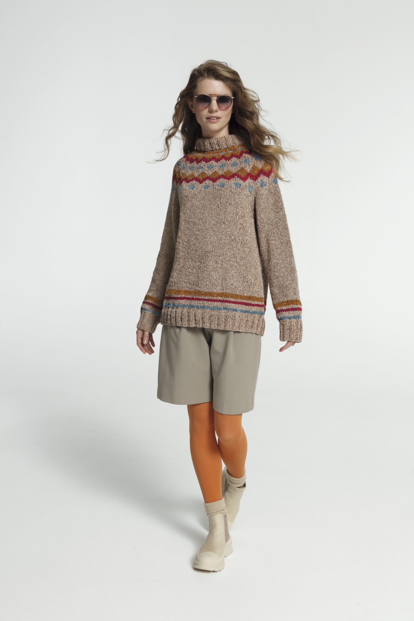 Modischer, handgestrickter Pullover mit buntem Muster, braune Shorts, orange Strumpfhose, cremefarbene Stiefel.