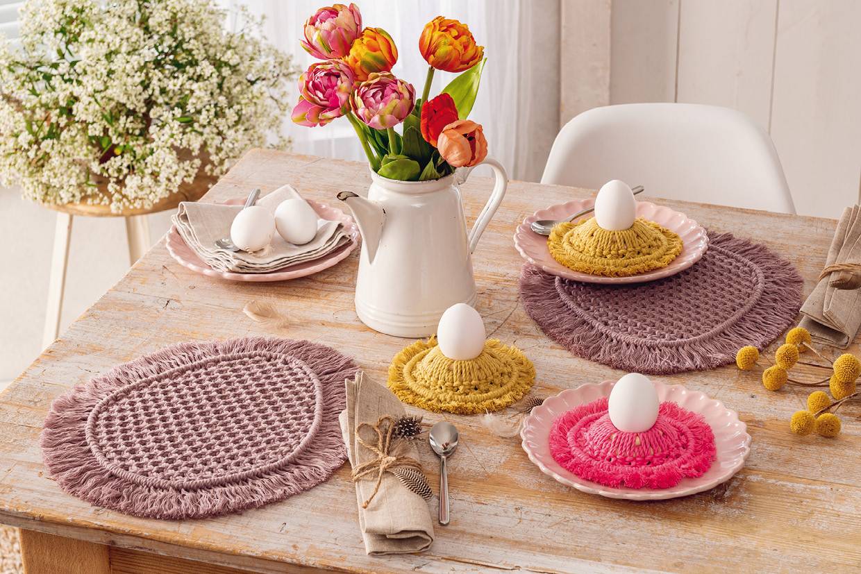Farbenfrohe, handgemachte Platzsets und Eierwärmer in Rosa und Gelb. Boho-Muster auf hellem Holztisch.