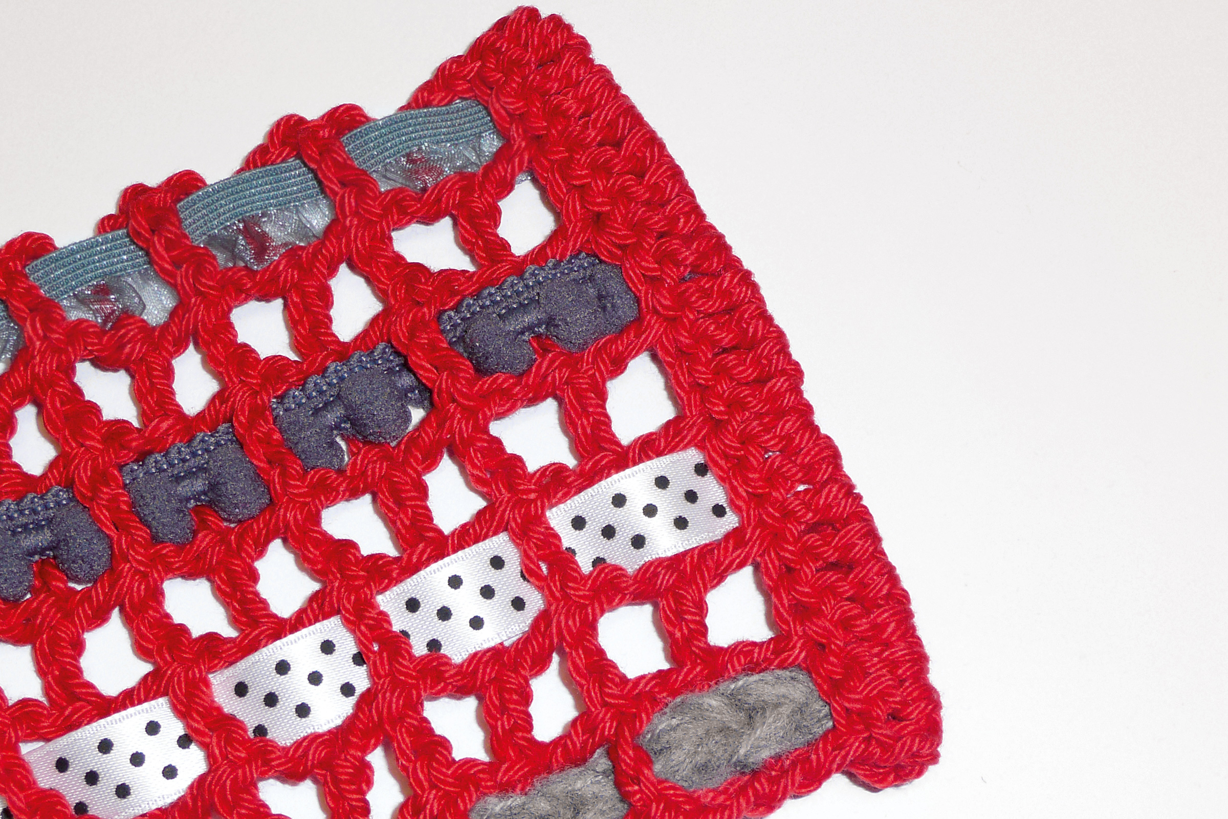Rotes, grobmaschiges, handgemachtes Häkelwerk mit eingearbeiteten Stoffstreifen in Blau und gepunktetem Weiß.