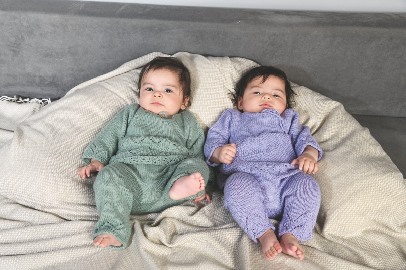 Zwei Babys in handgemachten Strickanzügen: grün und lila, mit zarten Musterdetails. Modische Babyoutfits.