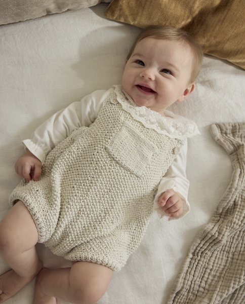 Cremefarbene, selbstgestrickte Baby-Latzhose mit feiner Struktur und Spitzenkragen; modische Handarbeit.