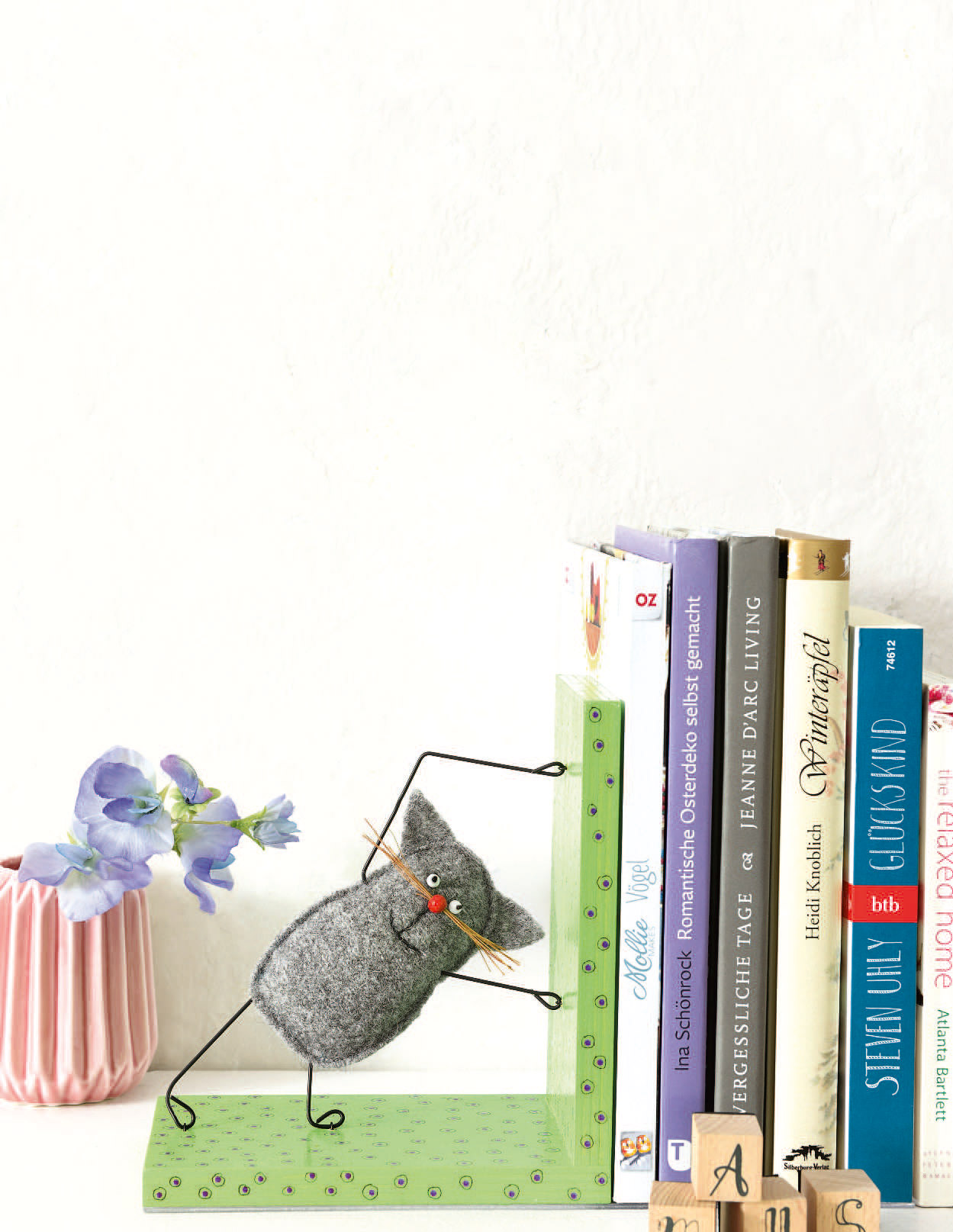 Graue Filzkatze mit roten Details, DIY-Stil, auf grüner Buchstütze, minimalistisch und kunstvoll gestaltet.