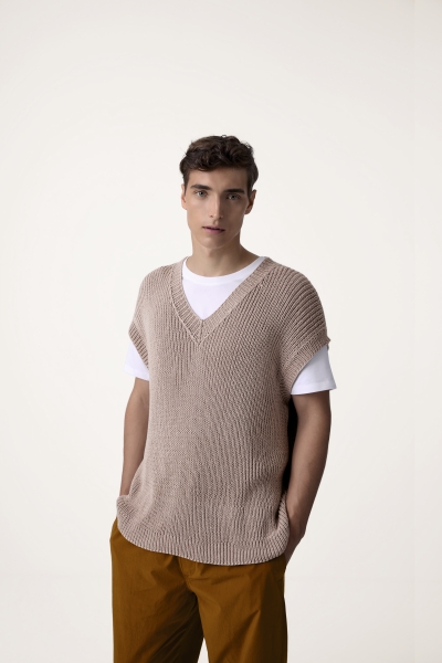 Beigefarbener, grob gestrickter Pullunder, V-Ausschnitt, über weißem Shirt, modern und handgemacht, kombiniert mit brauner Hose.