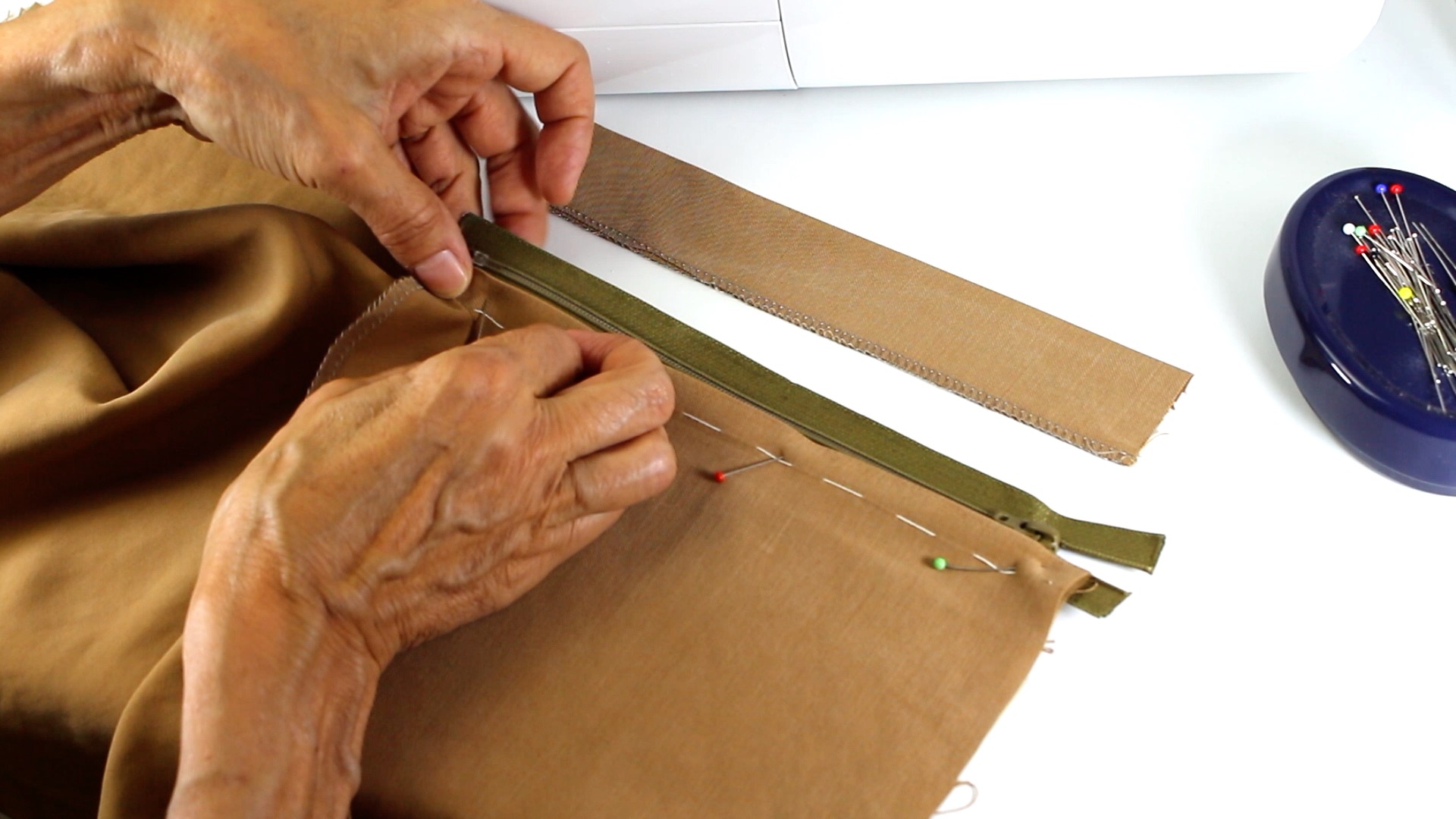 Brauner Stoff mit grünem Reißverschluss. Handgemacht, präzise Stichdetails, DIY, Nähnadeln sichtbar.