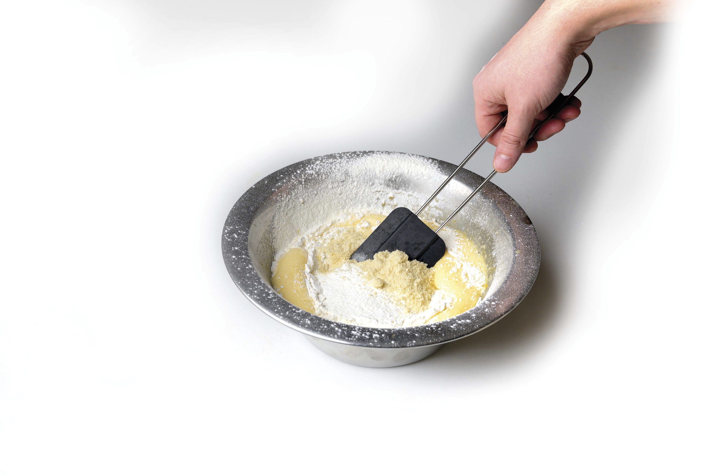 Hand mischt Zutaten in silberner Schüssel. Schwarzer Spatel, DIY-Backprozess, minimalistisch.