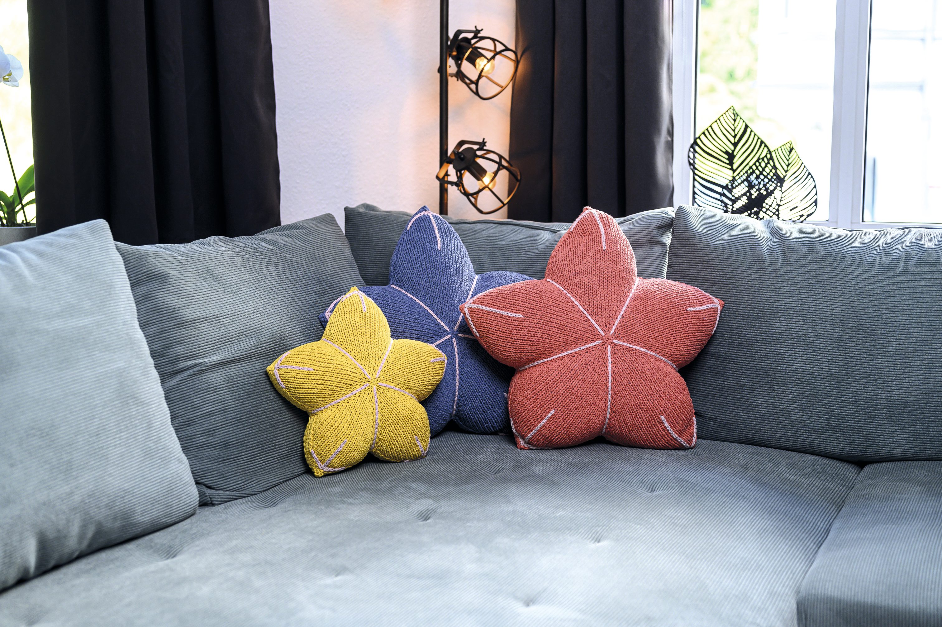 Drei bunte Blütenkissen in Gelb, Blau und Rot, handgemacht und modisch auf einem grauen Sofa arrangiert.