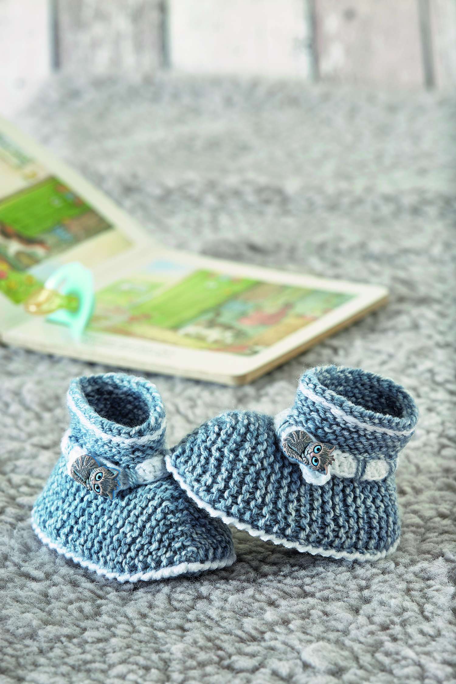 Blaue, gestrickte Babyschuhe mit weißer Borte und Knopfdetail. DIY, modisch.