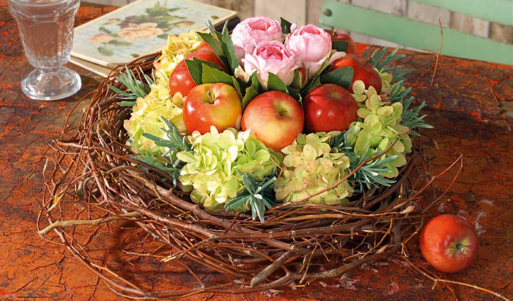 Erdfarbener DIY-Kranz, handgeflochten, dekoriert mit roten Äpfeln und rosa Blüten. Elegant und rustikal.