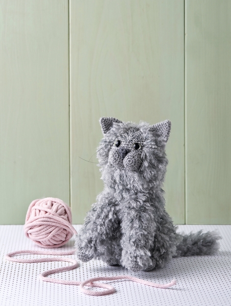 Graue, handgemachte Häkelkatze mit flauschigem Fell. Neben rosa Wollknäuel. Modisches DIY-Spielzeug.