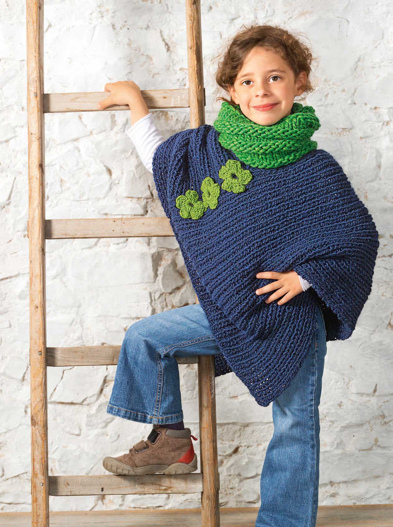 Blauer Poncho mit strukturierter Strickart, grünen Blüten und handgemachtem Charme; kombiniert mit blauer Jeans.