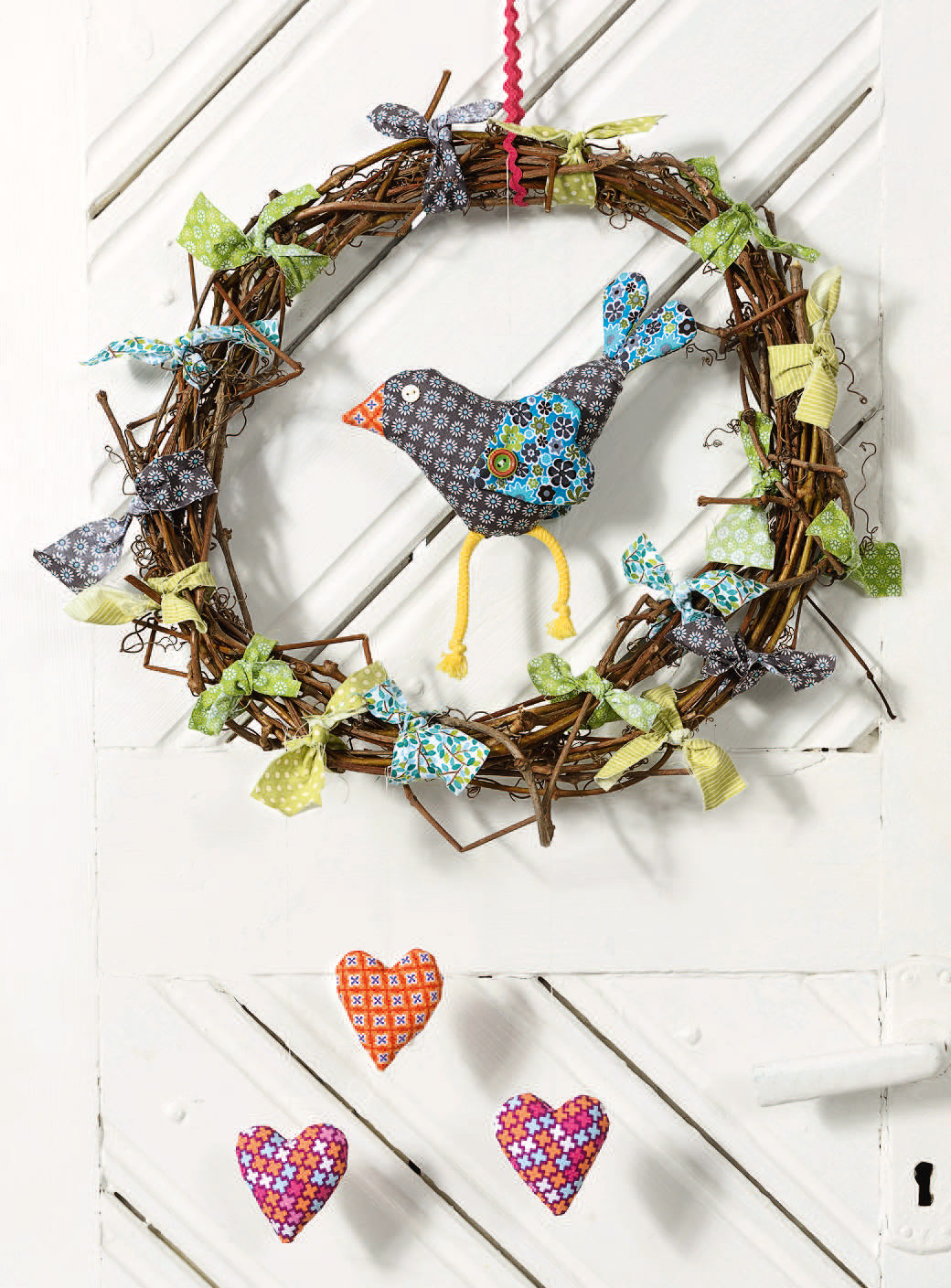 Handgemachter Kranz mit buntem Stoffvogel auf Zweigen, dekoriert mit farbenfrohen Schleifen und kleinen Herzanhängern.