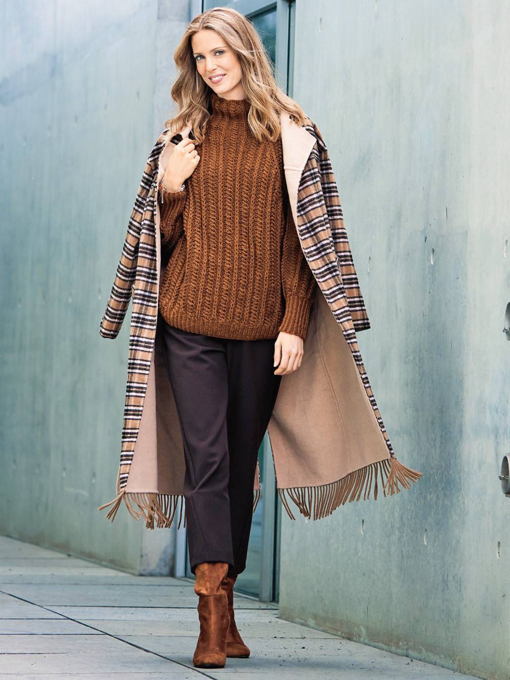 Geflochtener, brauner Pullover, karierter Mantel mit Fransen, schwarze Hose, passende Stiefel. Warm, stilvoll.