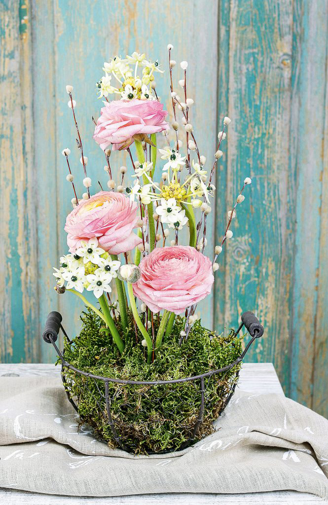 Zartes, handgemachtes Arrangement aus rosa Ranunkeln, weißen Blüten und Weidenkätzchen in einem Drahtkorb.