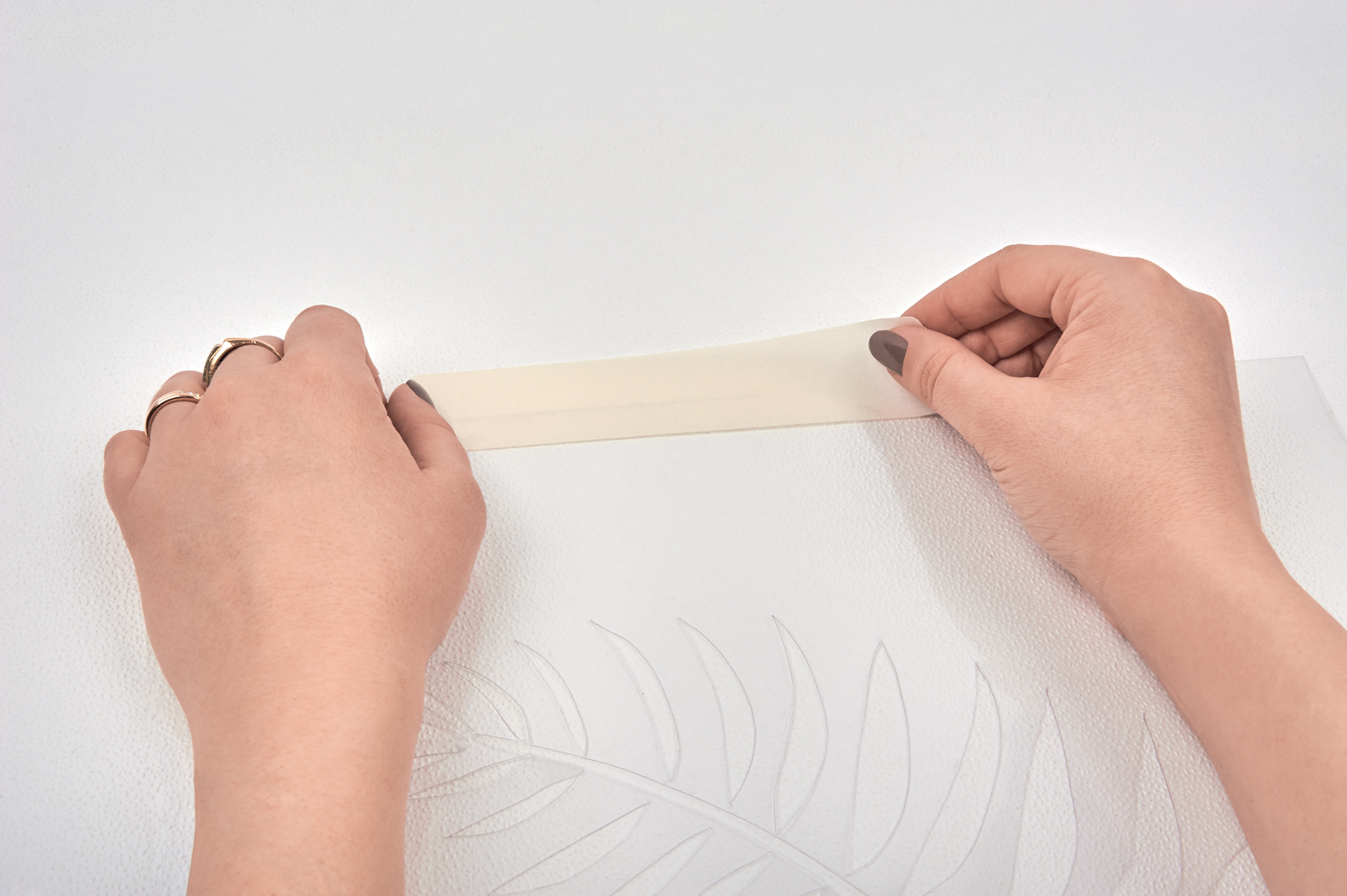 Zwei Hände kleben cremefarbenes Klebeband auf weiße Oberfläche mit Blattmuster. Handgemacht und elegant.