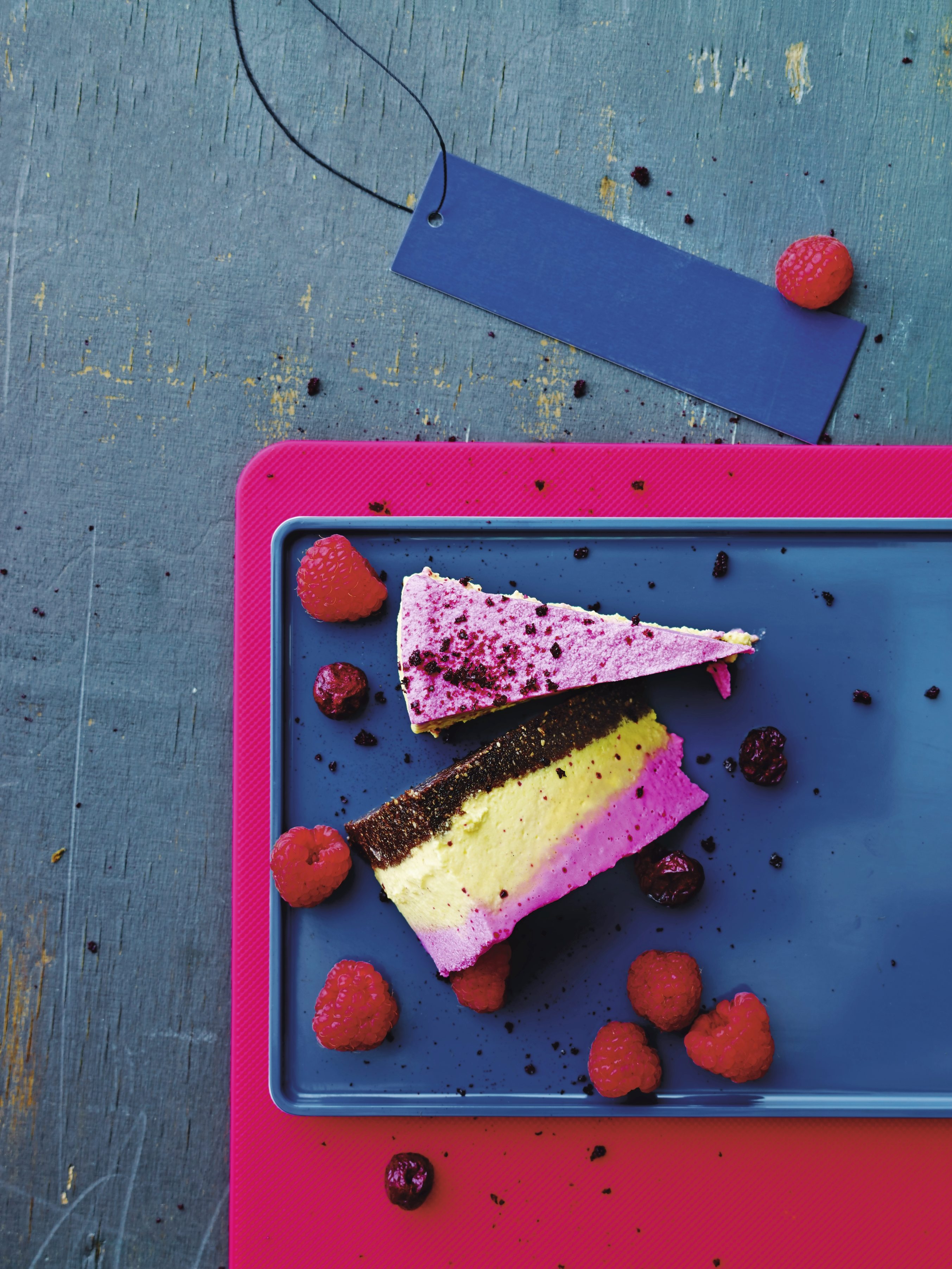 Bunte, handgemachte Kuchenstücke in Gelb und Pink, garniert mit Himbeeren auf blauem Teller. Ein echter Farbklecks.