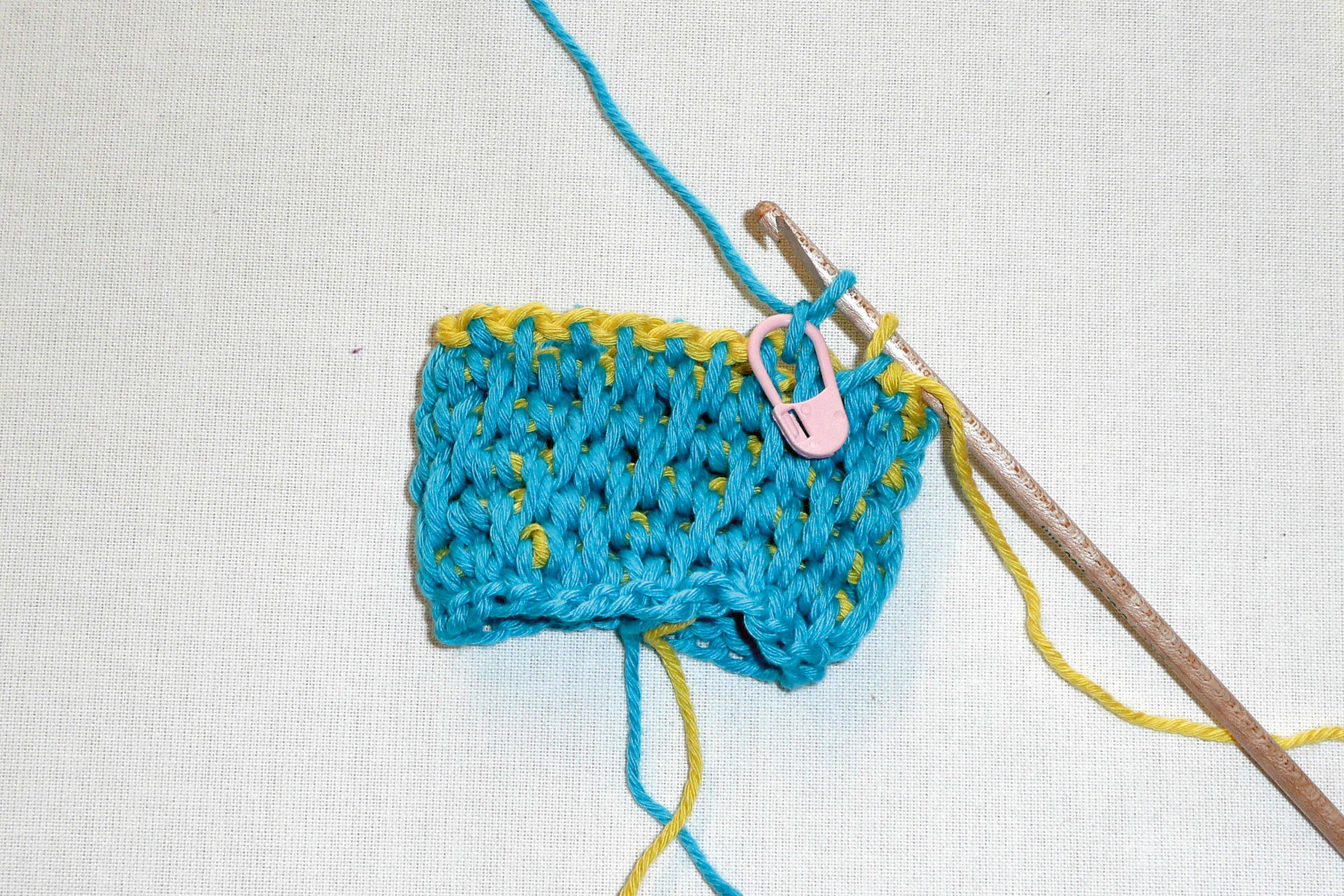 Buntes, selbstgemachtes Strickstück in Blau und Gelb. Modische Handarbeit mit Häkelhaken und pinker Maschenmarkierung.