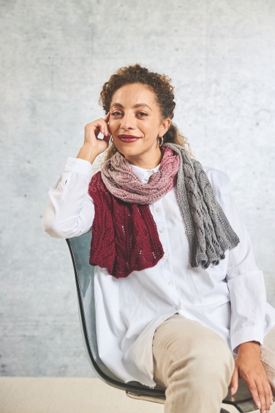 Farbenfrohe, handgefertigte Strickschals in Grau, Weinrot und Rosa, kombiniert zu schlichter weißer Bluse.