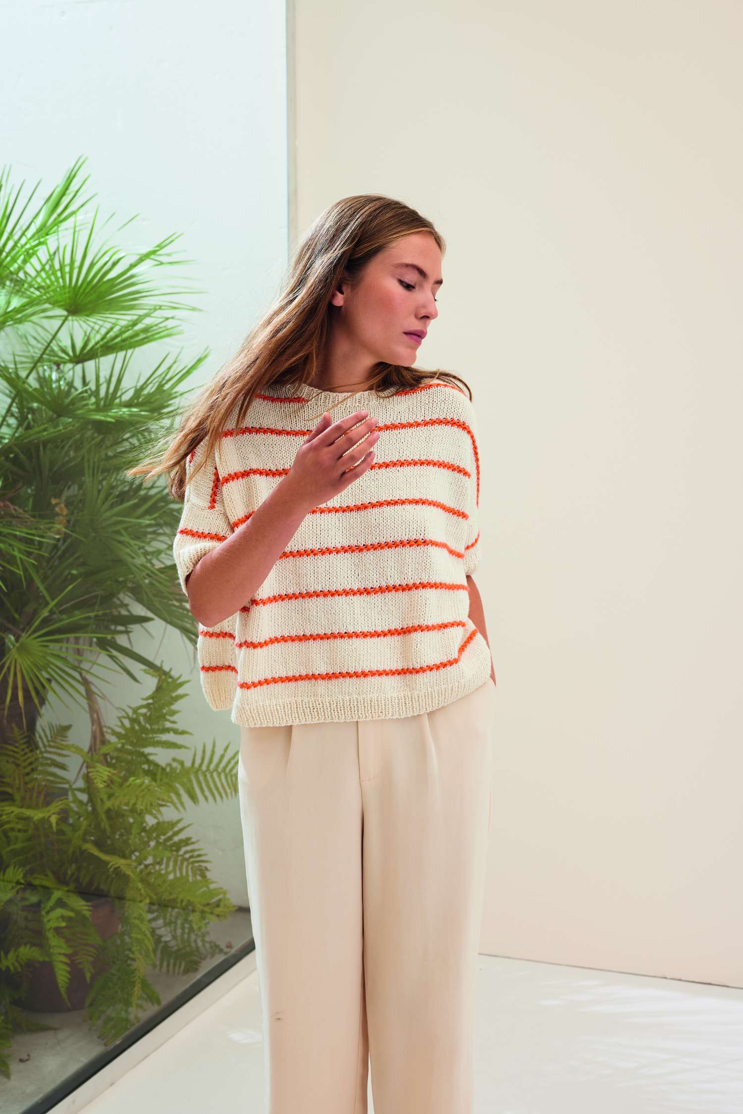 Cremefarbener Strickpullover mit orangen Streifen, kurzarm, kombiniert mit legeren, hellen Hosen. Modisch, handgearbeitet.