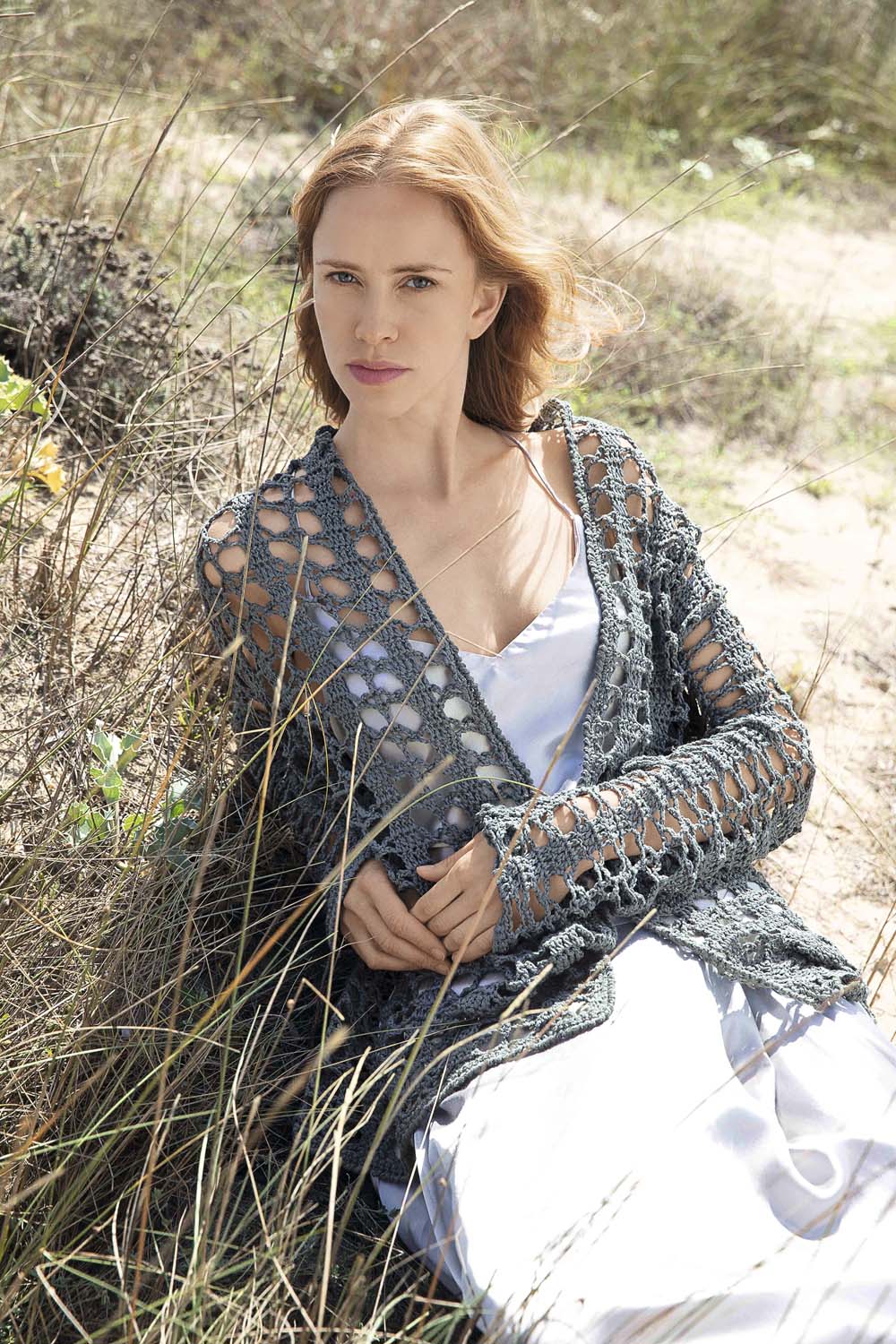 Graue, gehäkelte Weste, offen und luftdurchlässig, über weißem Kleid. Stilvoll, handgemacht, ideal für den Sommer.