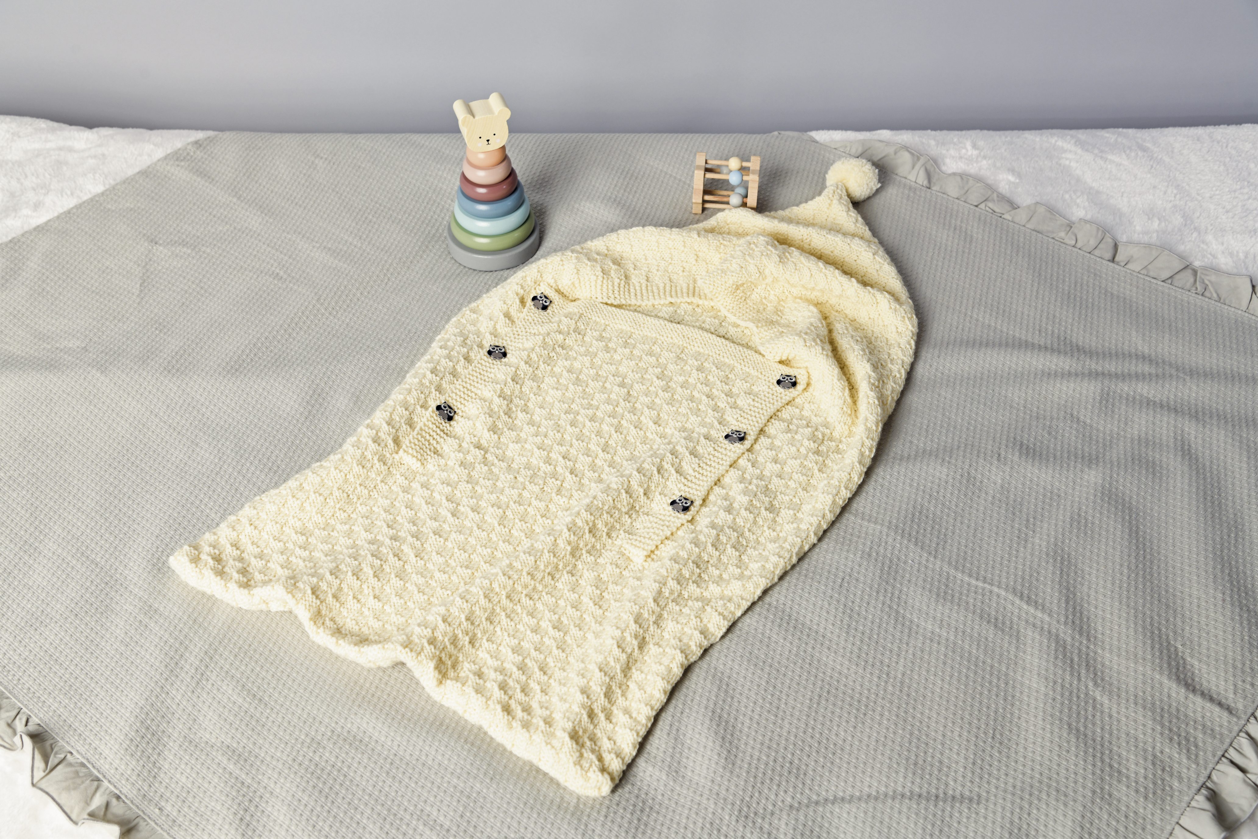 Handgemachte cremefarbene Babydecke mit Pom-Pom und Knopfdetails. Strickmuster: zart, texturiert.