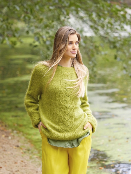 Ein moosgrüner, handgemachter Strickpullover mit Zopfmuster, kombiniert mit leuchtend gelben, lockeren Hosen.