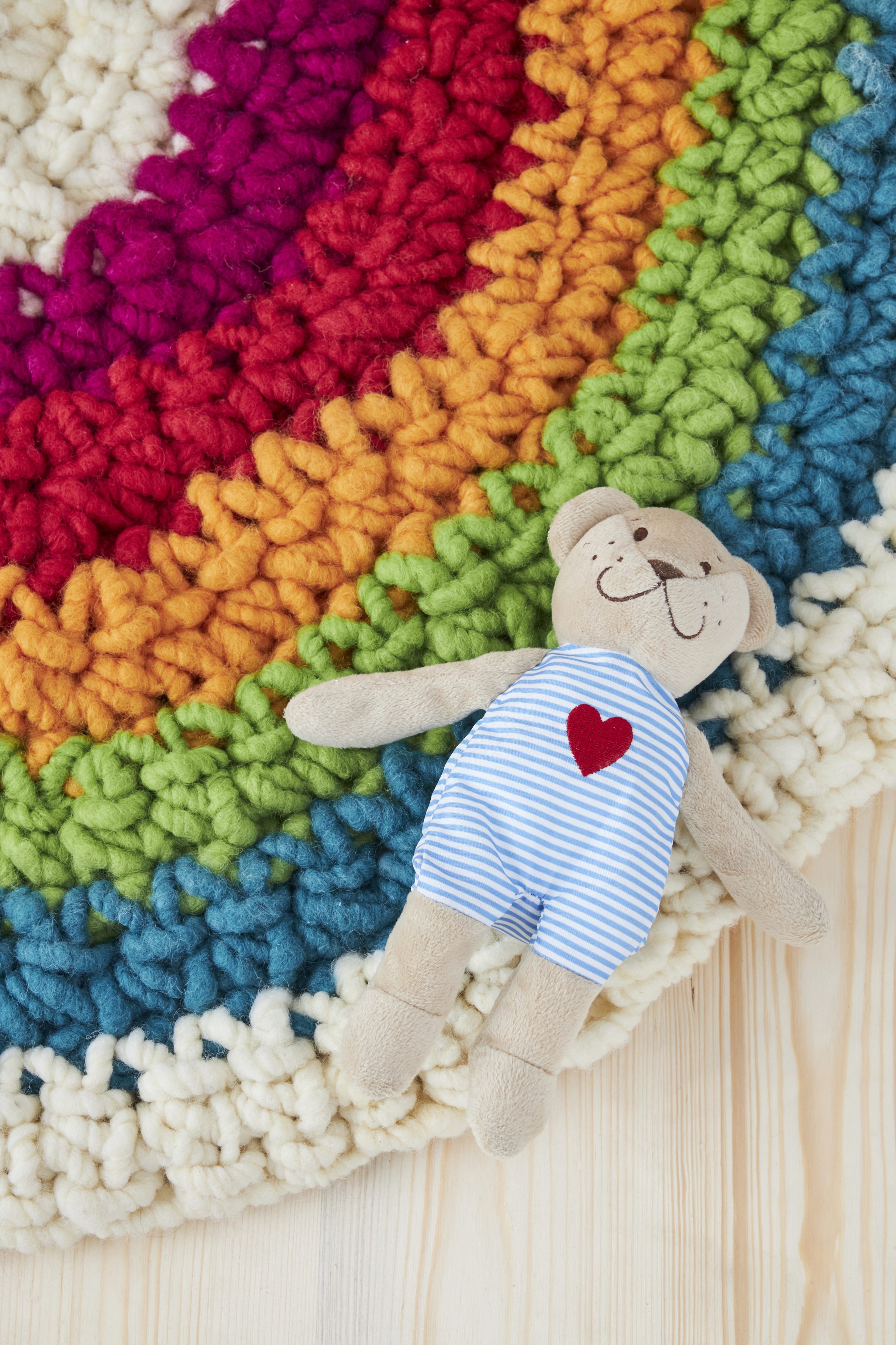 Puppe in gestreiftem Overall mit rotem Herz, auf handgemachtem Regenbogen aus bunter Wolle.